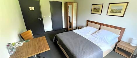 A hotel room with a bed, bedside table, chair, and a desk with a chair. There are two doors, one with a 'No Entry' sign, and two framed pictures on the wall.