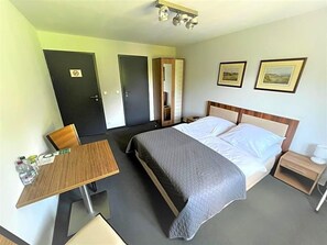 A hotel room with a bed, bedside table, chair, and a desk with a chair. There are two doors, one with a 'No Entry' sign, and two framed pictures on the wall.