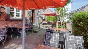 An outdoor patio area with wicker furniture and red umbrellas, featuring a brick wall and a view of the street. - Hotel Restaurant Mathildenhof (Pulheim)