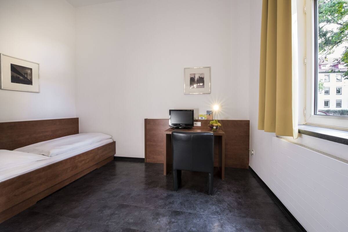 A hotel room with a single bed, a desk with a computer, and a window with curtains.