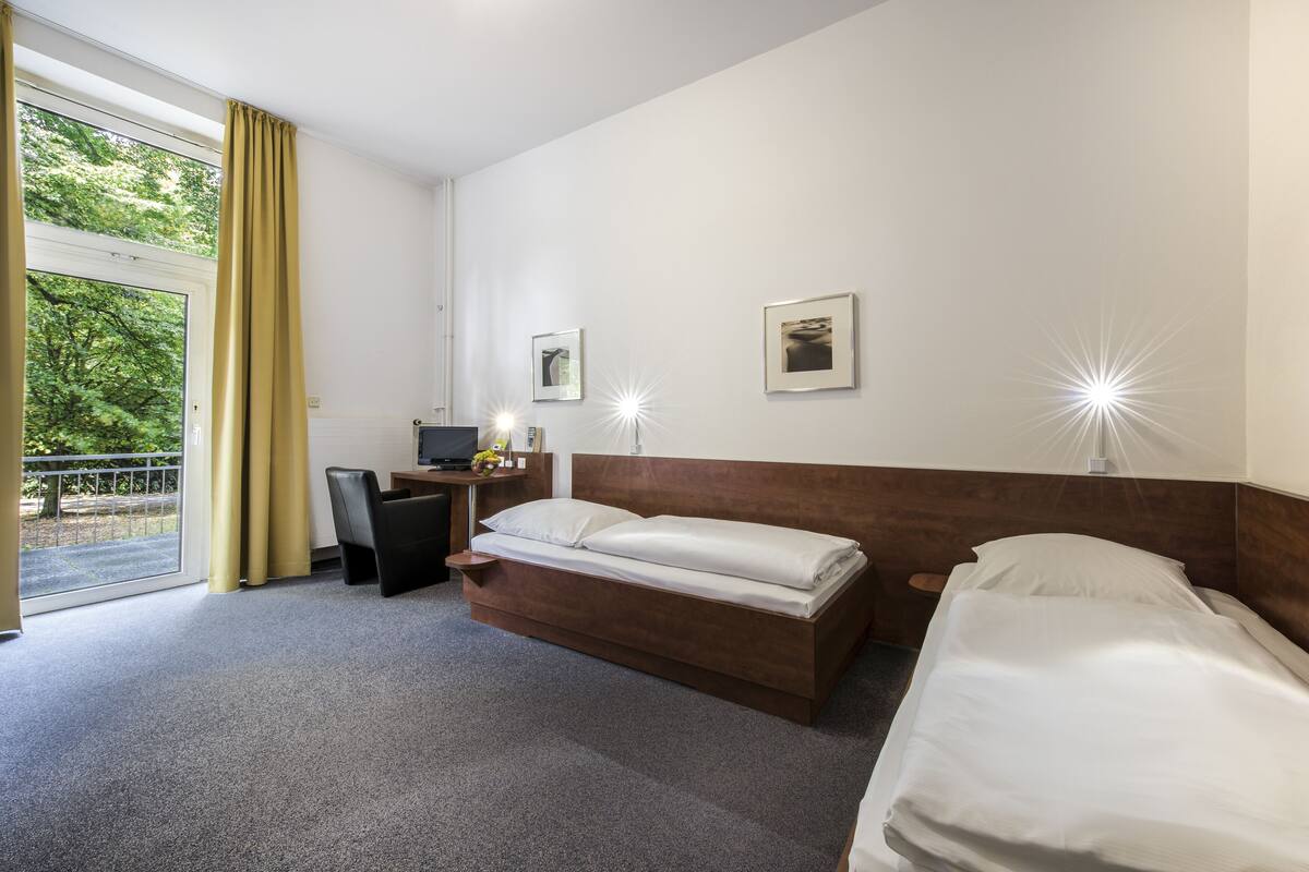 A hotel room with a bed, a desk, and a chair. There are two bedside lamps and a window with curtains.
