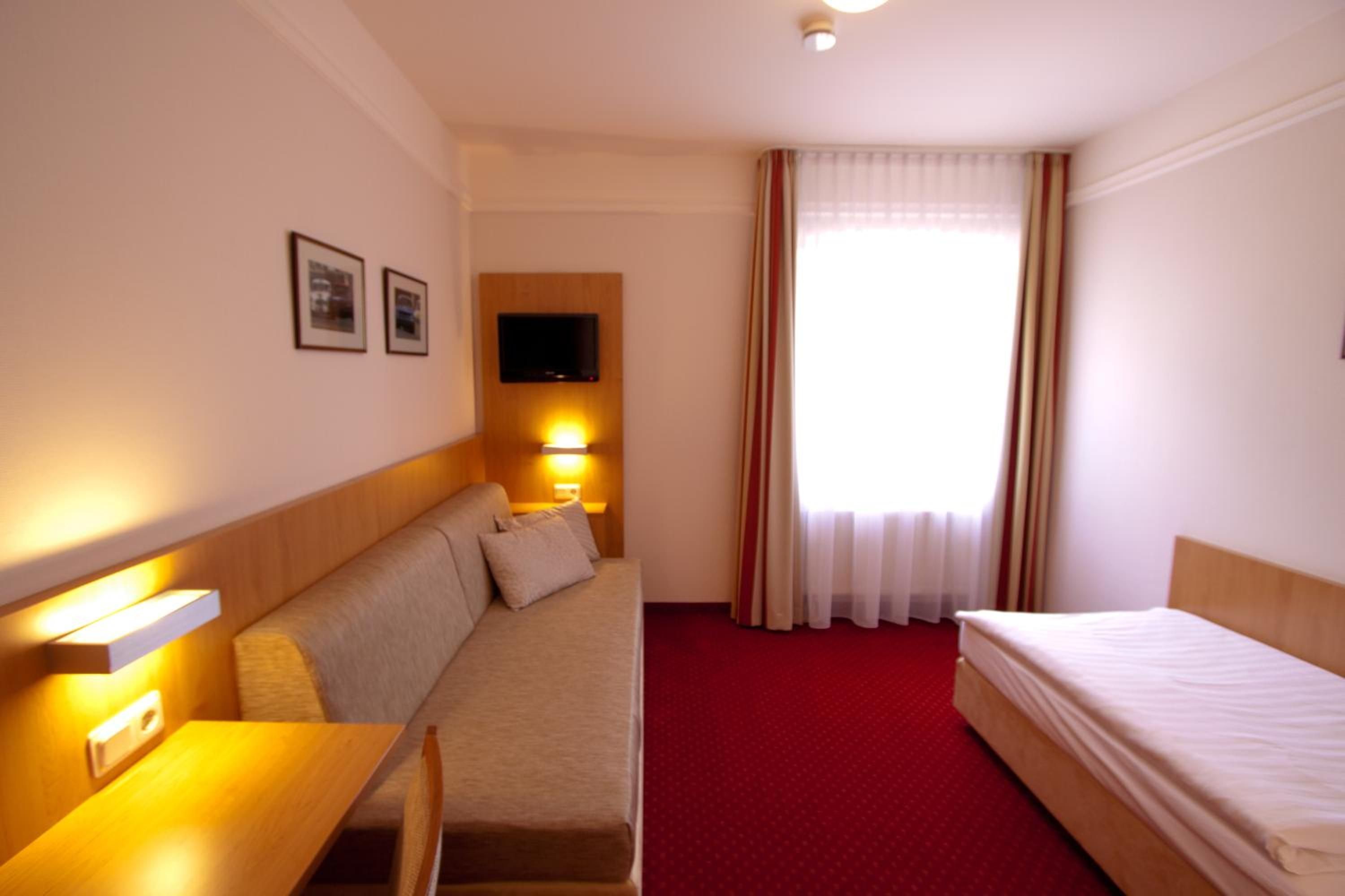 A hotel room with a sofa, a bed, a desk, a television mounted on the wall, and a window with curtains.