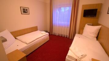 Ein Hotelzimmer mit zwei Einzelbetten, einem an der Wand befestigten Fernseher und einem Fenster mit durchscheinenden Vorhängen.