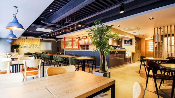 Um café moderno com mesas e cadeiras de madeira, um balcão e uma planta grande no centro.