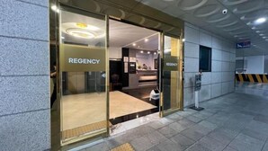 Entrada de um hotel com uma recepção, uma placa indicando "REGENCY" e uma câmera de segurança no teto.