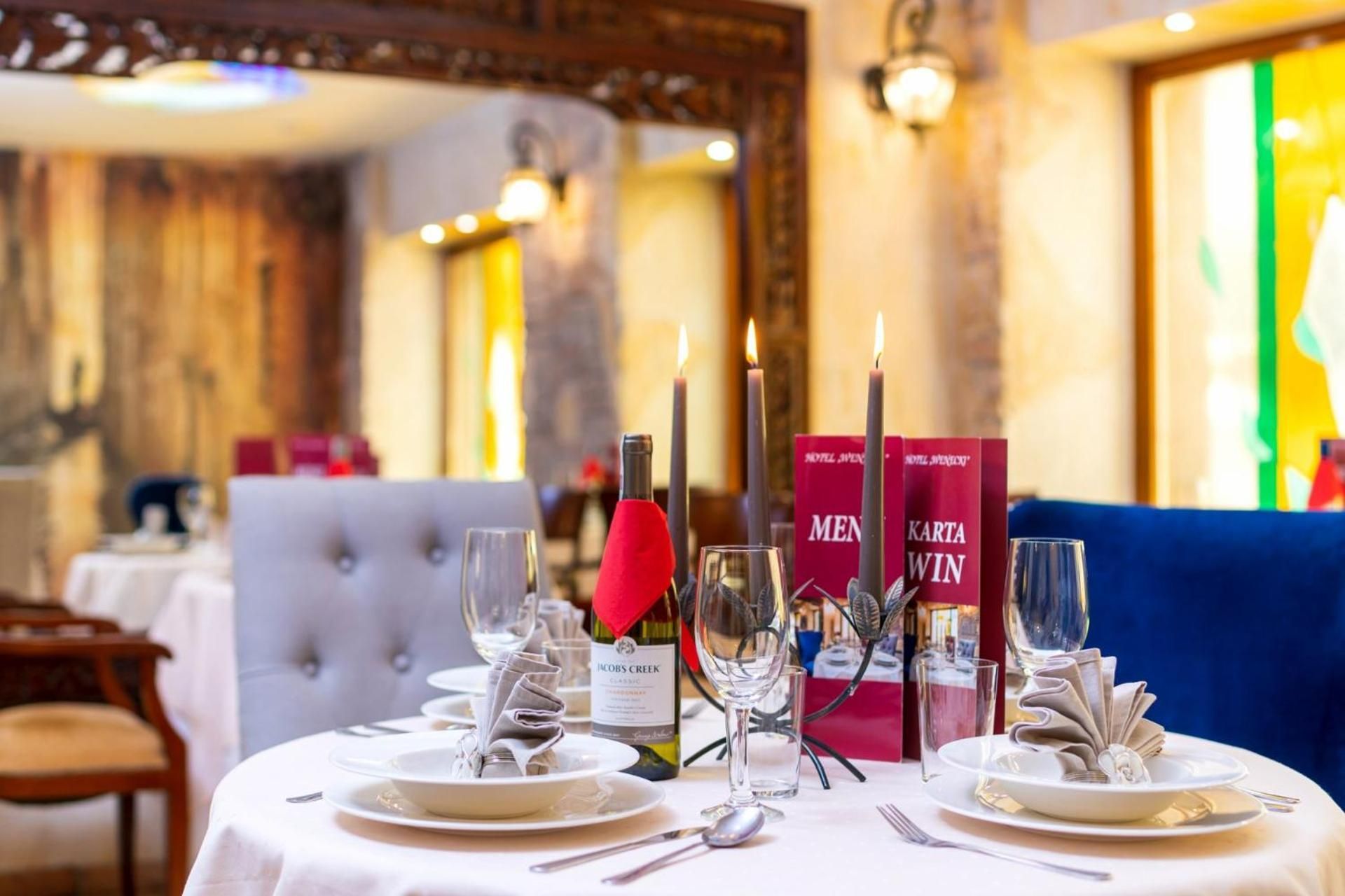 Una mesa de comedor dispuesta con platos, cubiertos, copas y velas, con libros y una botella de vino sobre la mesa.