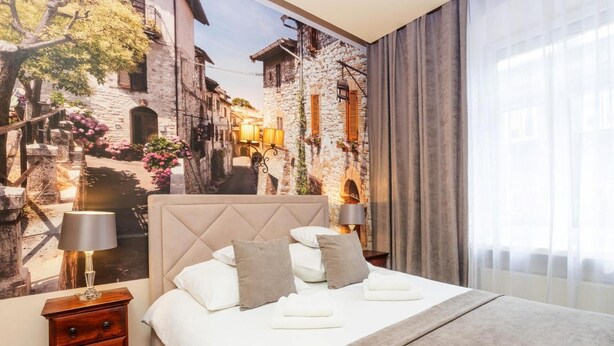 Een hotelkamer met een bed, nachtkastje, lamp en uitzicht op een straat.