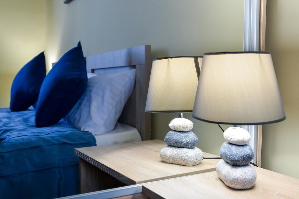 A neatly made bed with blue bedding and pillows, a bedside table with two lamps featuring stacked stones, and a mirror reflecting the room's interior.