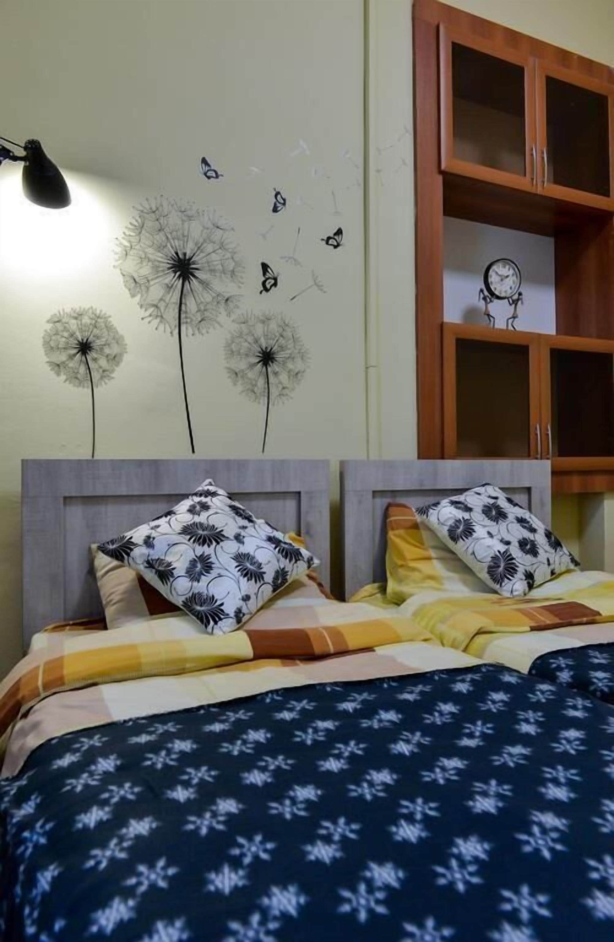 A bed with floral-patterned pillows, a dandelion wall decal, and a clock on the wall.