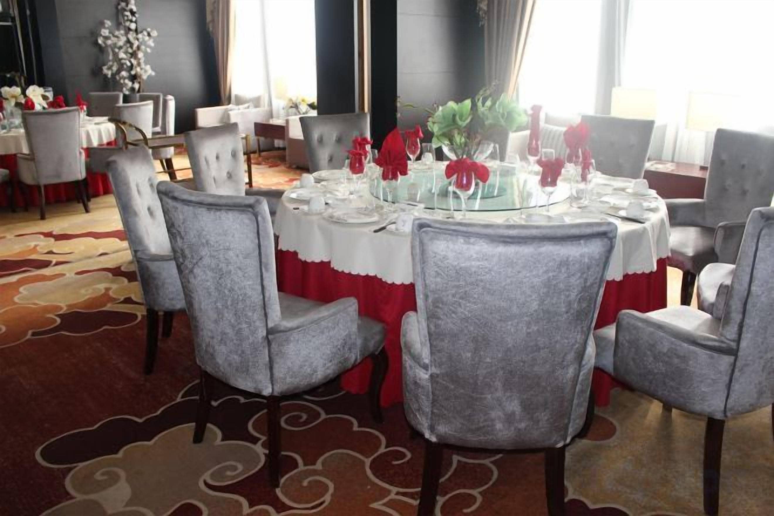 A round dining table with grey upholstered chairs, set with white tablecloths and red accents, in a room with patterned carpet and large windows.