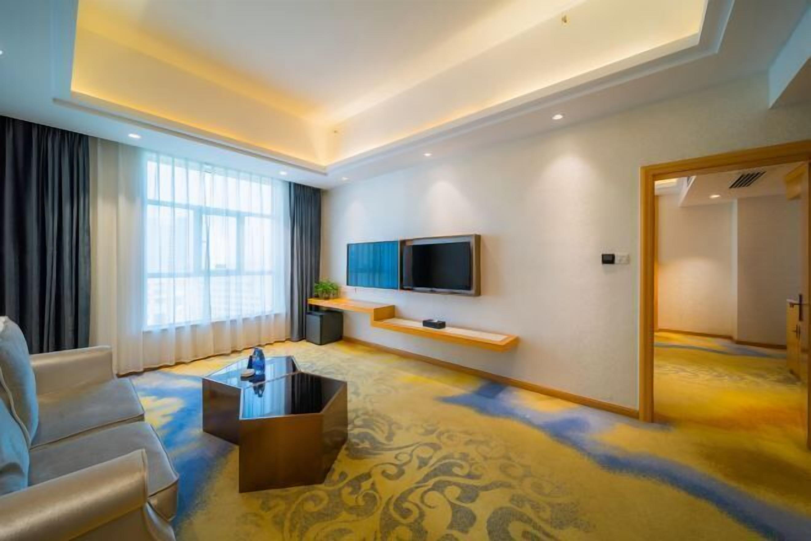 Ein modernes Hotelzimmer mit einer Couch, einem Couchtisch, einem an der Wand befestigten Fernseher und einem gemusterten Teppich.