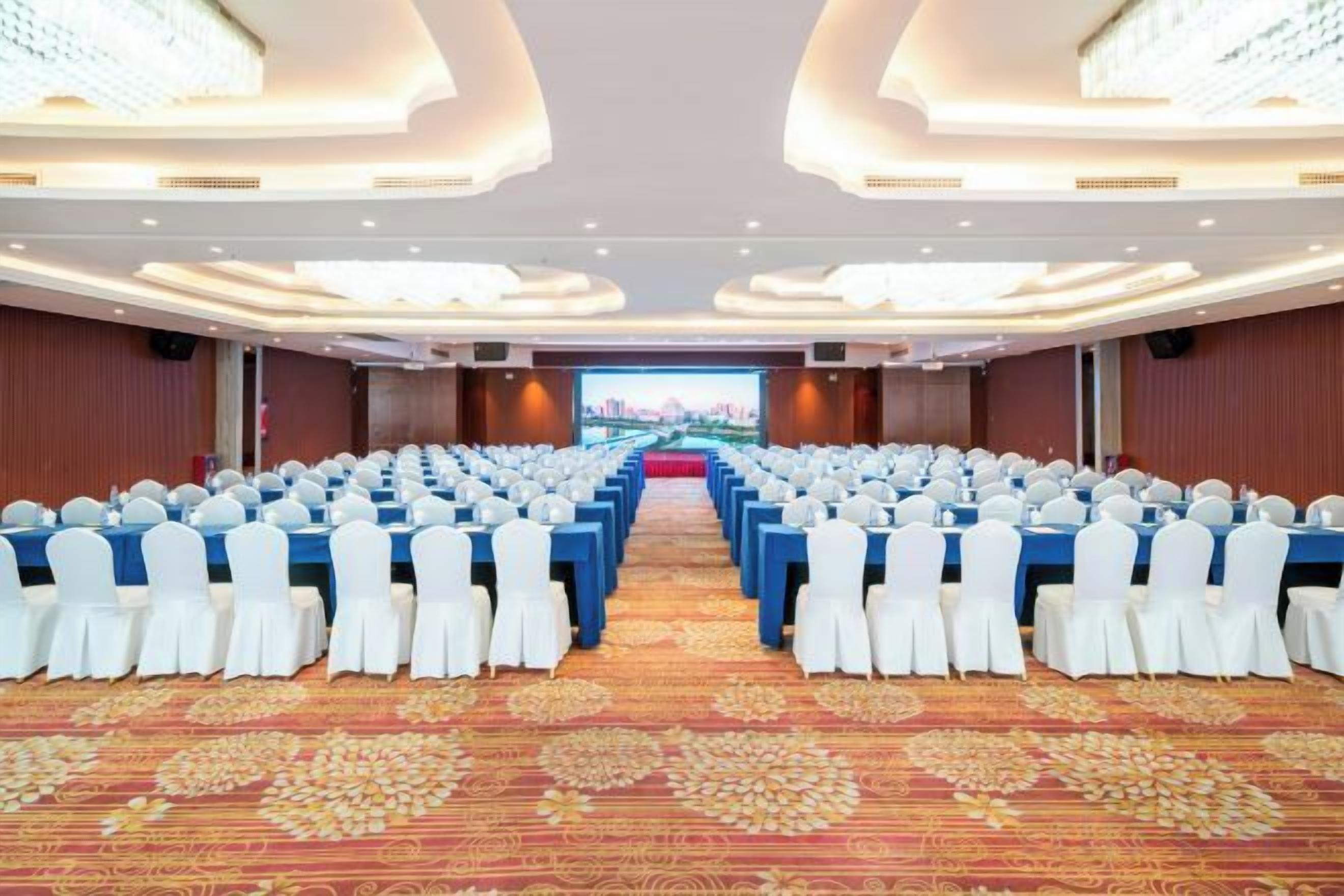 Un salón de conferencias con filas de mesas y sillas, piso alfombrado y una pantalla grande al frente.