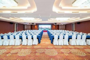Un salón de conferencias con filas de mesas y sillas, piso alfombrado y una pantalla grande al frente.