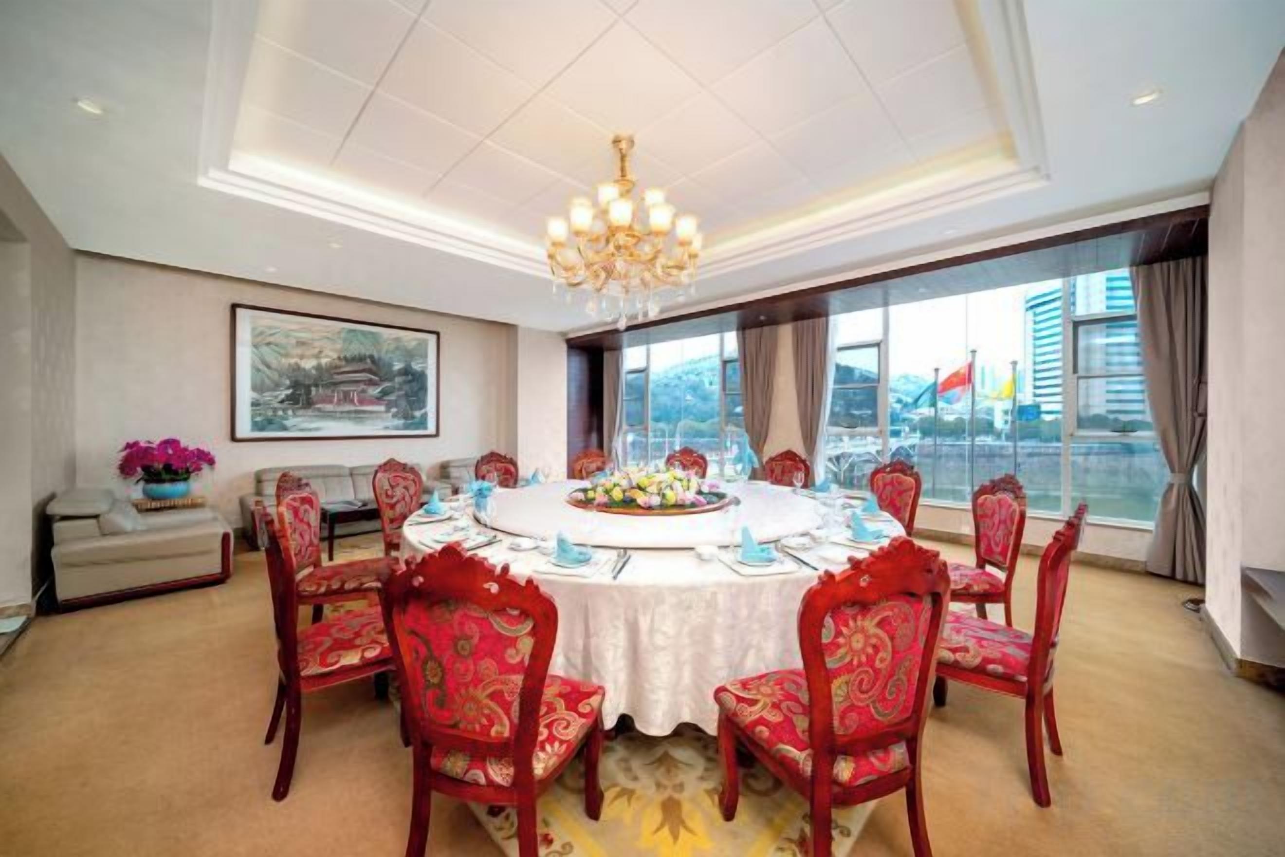 Uma sala de conferências com uma mesa redonda, cadeiras estofadas em vermelho, um candelabro e uma grande janela com vista para a cidade.