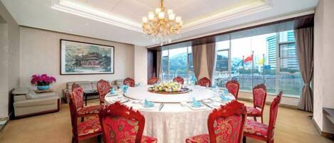Uma sala de conferências com uma mesa redonda, cadeiras estofadas em vermelho, um candelabro e uma grande janela com vista para a cidade.