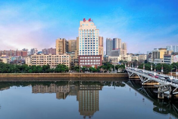 A cityscape with a prominent building reflecting on calm water, a bridge, and a clear sky.