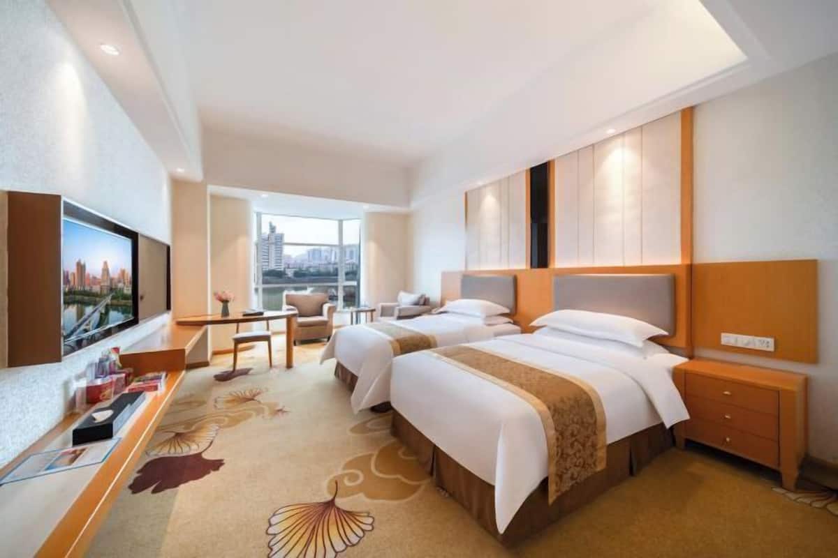 A hotel room with two beds, a desk, a TV, and a view of a cityscape.