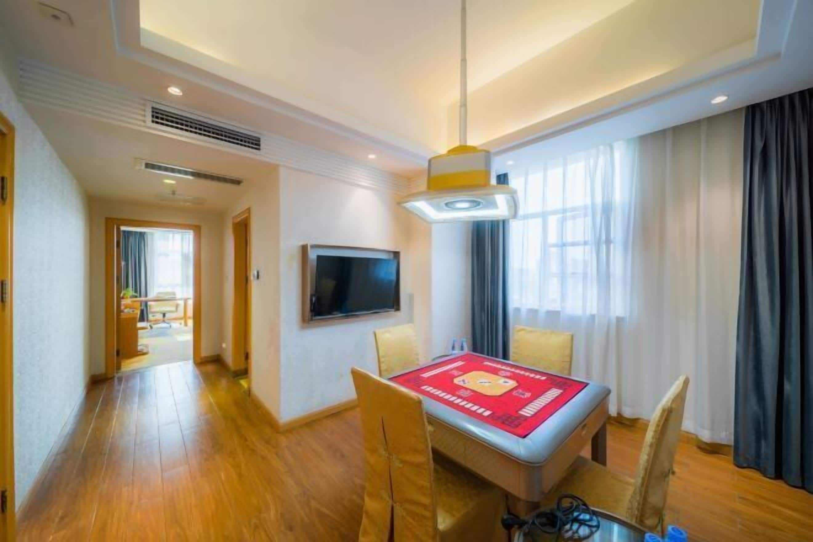 Quarto de hotel com piso de madeira, mesa de jantar com cadeiras, TV de tela plana na parede e uma janela grande com cortinas.