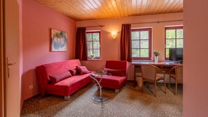 A cozy room with a red sofa, a small table, and a desk with a chair. There are windows with curtains, a painting on the wall, and a ceiling with wooden beams. - Berggasthof Butterberg (Bischofswerda)