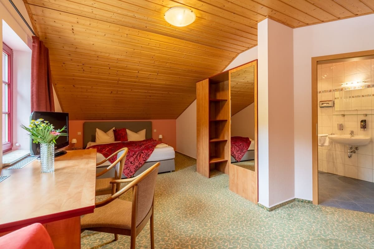 A bedroom with a bed, a desk, a chair, a TV, and a bathroom with a sink and mirror.