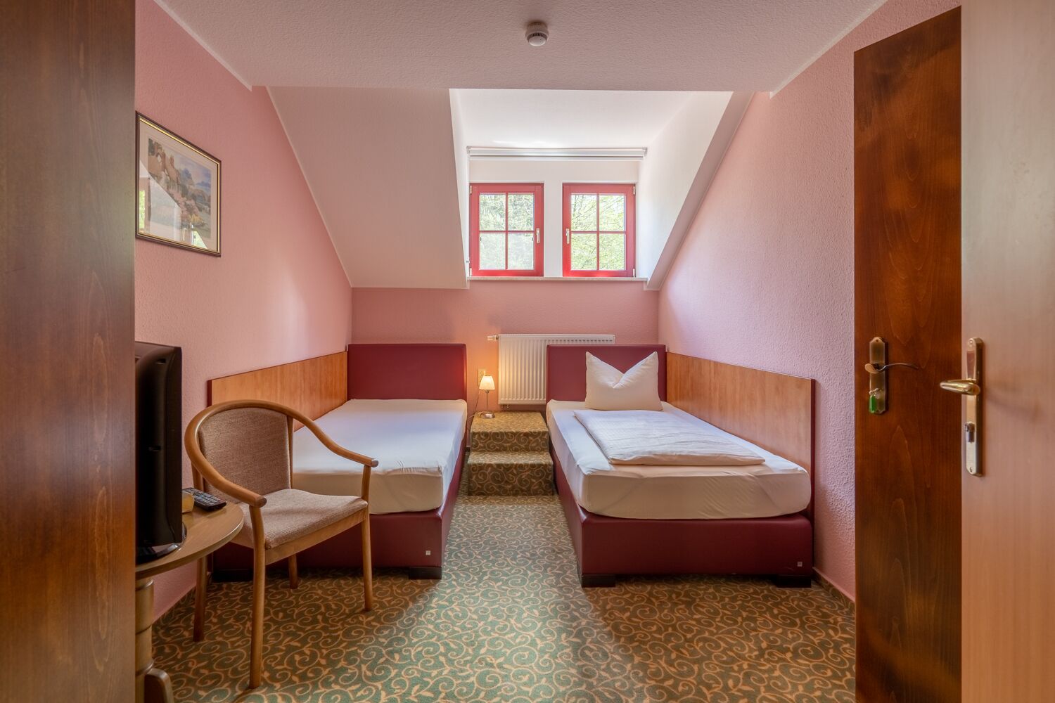 Una habitación pequeña y acogedora con dos camas individuales, una silla, un escritorio y una ventana con contraventanas rojas.
