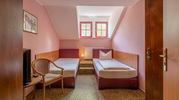 Una habitación pequeña y acogedora con dos camas individuales, una silla, un escritorio y una ventana con contraventanas rojas.