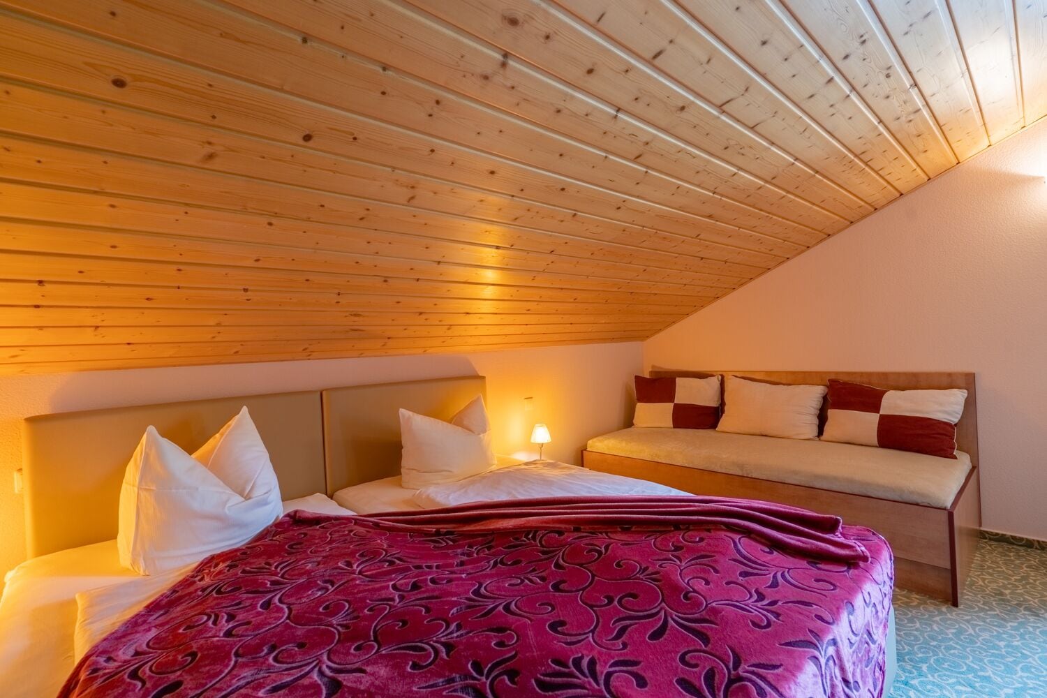 Ein Schlafzimmer mit geneigter Decke, einem Bett mit rotem Muster und einem Nachttisch mit Lampe.