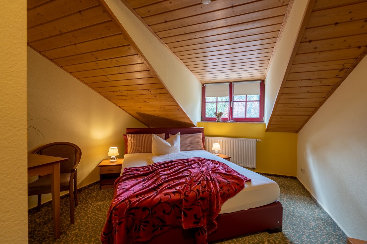 A cozy attic bedroom with a wooden ceiling, a single bed with a red blanket, a nightstand with a lamp, and a window with a view of trees.