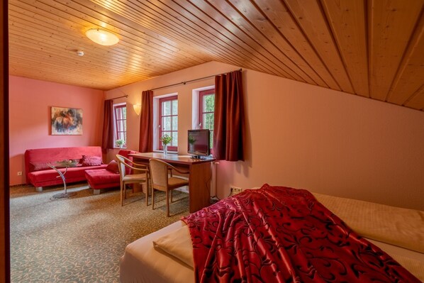 Une pièce avec un plafond en bois, un lit recouvert d’une couverture à motifs rouges, un bureau avec un ordinateur, deux chaises, un canapé rouge et un tableau accroché au mur.