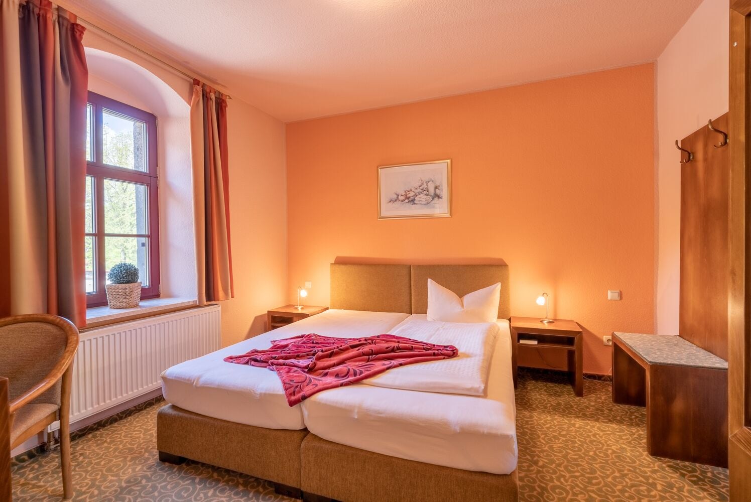 Habitación de hotel con cama doble, dos mesitas de noche, una silla, ventana con cortinas, radiador y un cuadro en la pared.
