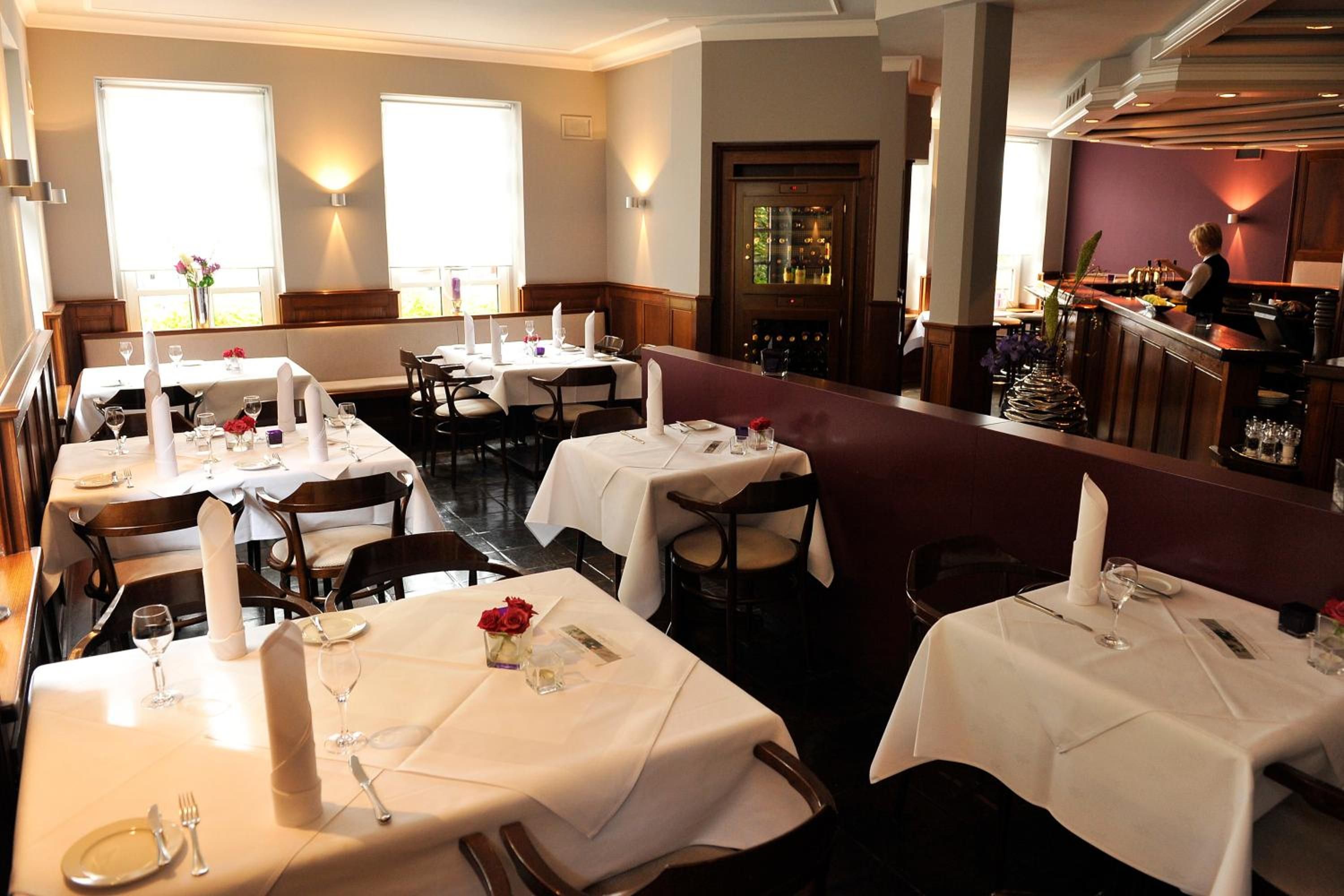 Ein Restaurant mit mehreren Tischen, einer Bar und Blick ins Freie durch große Fenster.