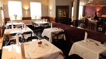 Ein Restaurant mit mehreren Tischen, einer Bar und Blick ins Freie durch große Fenster.