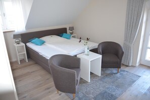 A modern bedroom with a large bed, two bedside tables, a small table, two armchairs, and a window with curtains.