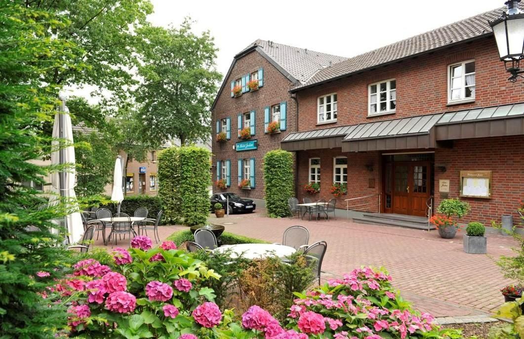 Ein Backsteinhaus mit überdachter Außenbestuhlung, einem davor geparkten Auto und einem Garten mit rosa Blumen.