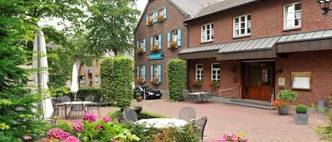 Un bâtiment en briques doté d’un espace extérieur couvert avec des sièges, une voiture garée devant et un jardin agrémenté de fleurs roses.