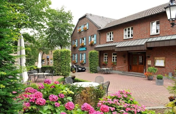 A brick building with a covered outdoor seating area, a car parked in front, and a garden with pink flowers.
