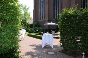 An outdoor patio area with tables and chairs, surrounded by greenery and a brick building in the background.