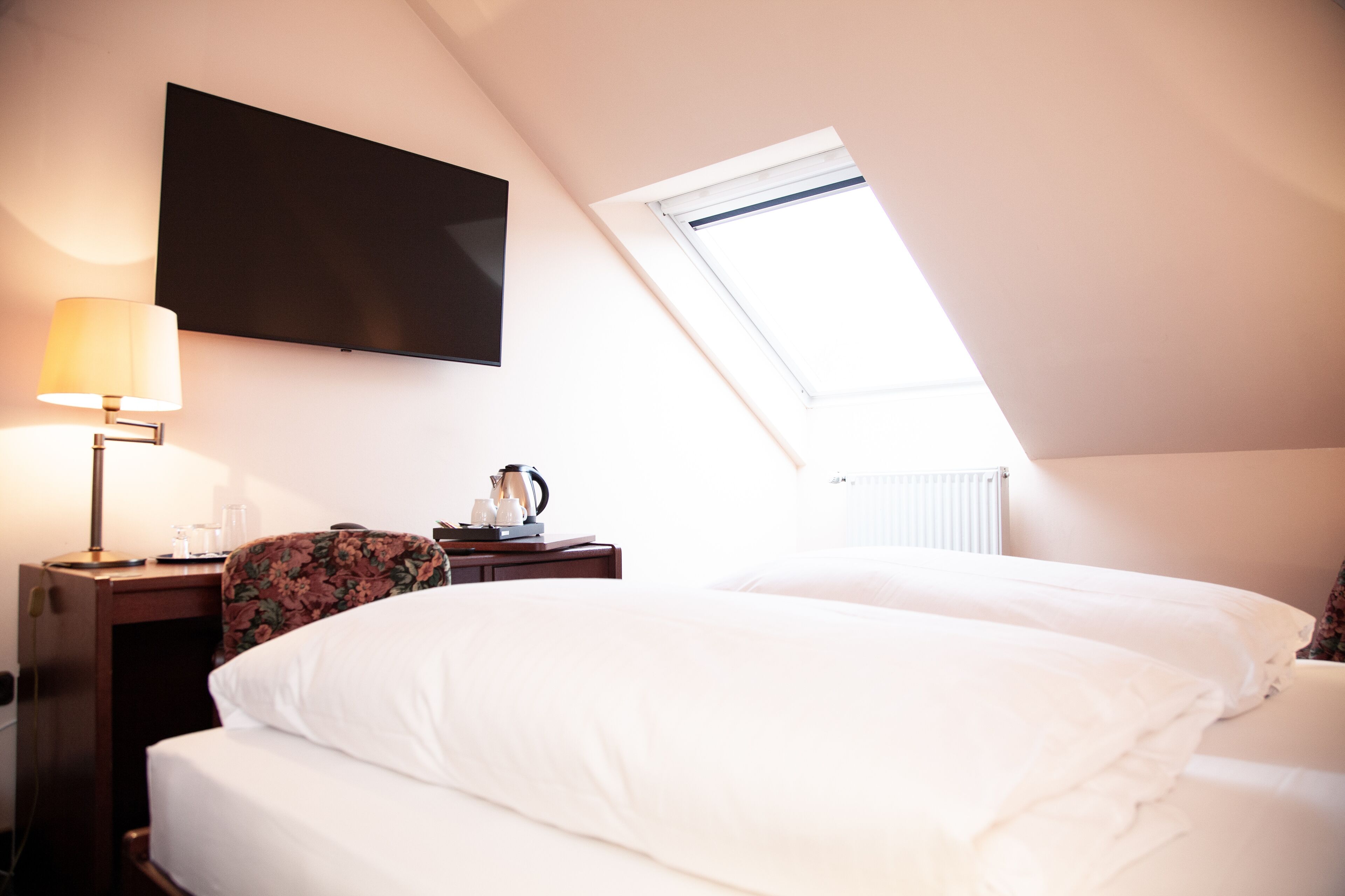 A bedroom with a bed, a desk with a lamp, a flat-screen TV, and a window.