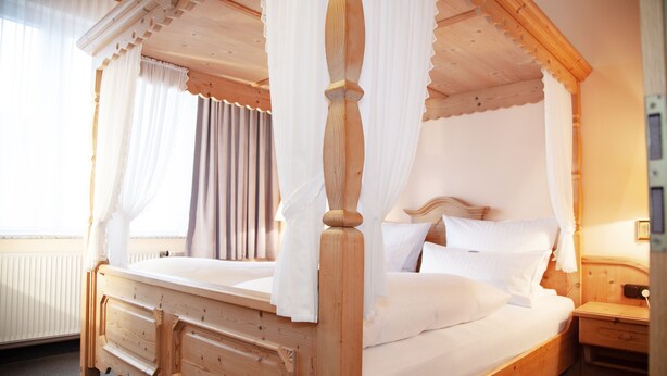 A wooden four-poster bed with white bedding, a wooden nightstand, and a window with curtains. - Hotel Lonac (Duisburg)