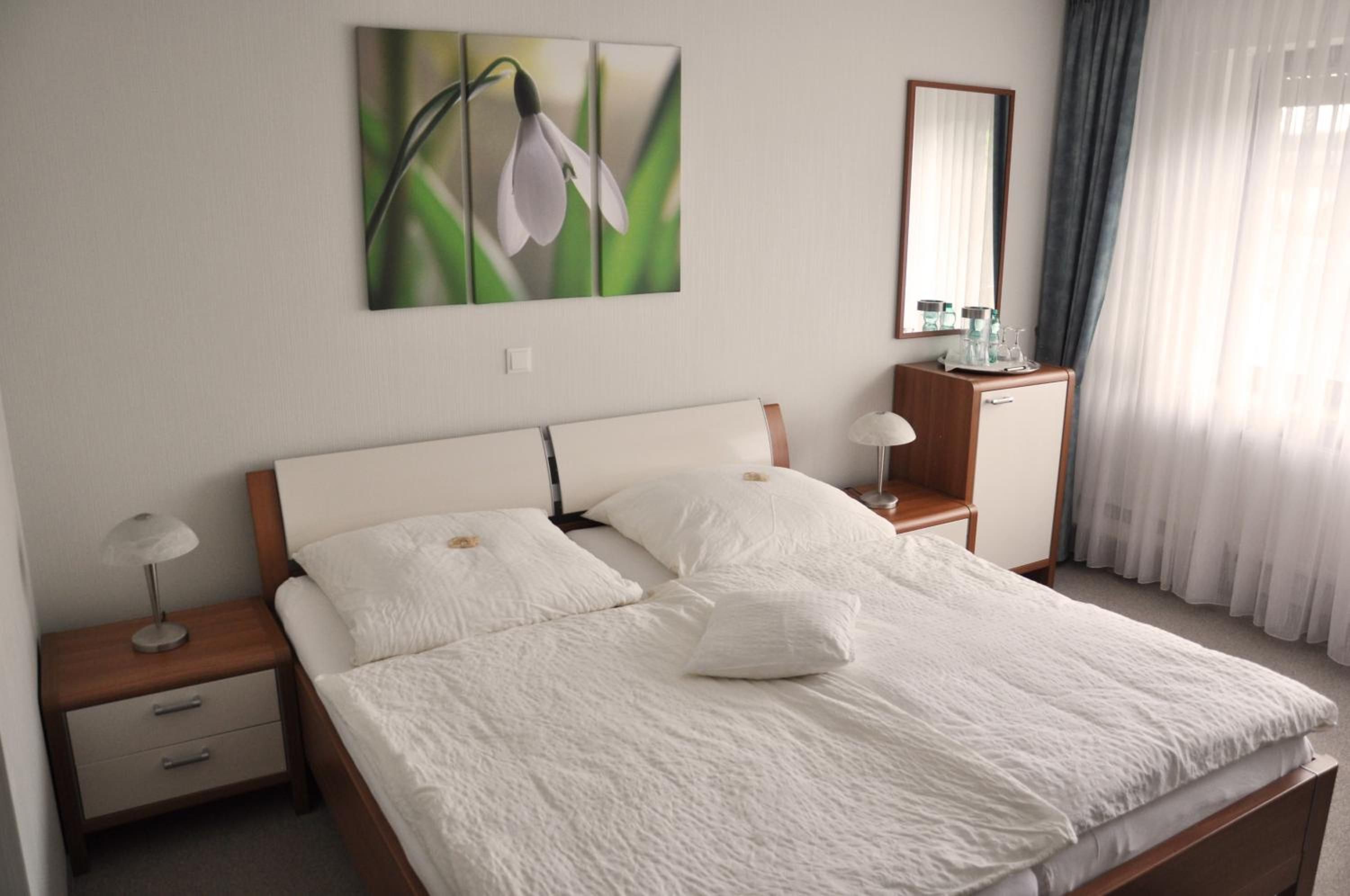 Ein ordentlich bezogenes Bett mit weißen Leinen und Kissen, ein Nachttisch mit Lampe, ein kleiner Holzbeistelltisch, ein Spiegel und ein Fenster mit durchsichtigen Vorhängen.