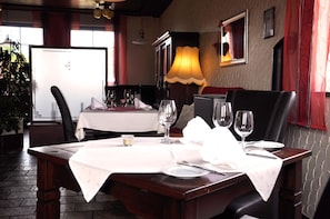 Une table à manger dressée avec des nappes blanches, des assiettes et des verres à vin, agrémentée d’une lampe et d’un tableau encadré au mur.