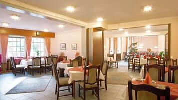 A hotel dining area with multiple tables and chairs, a large window with curtains, and a plant in the corner.