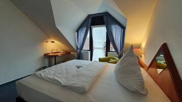 A hotel room with a bed, a small table, a chair, a window with curtains, and a lamp.