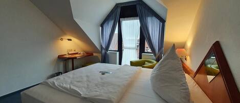 A hotel room with a bed, a small table, a chair, a window with curtains, and a lamp.