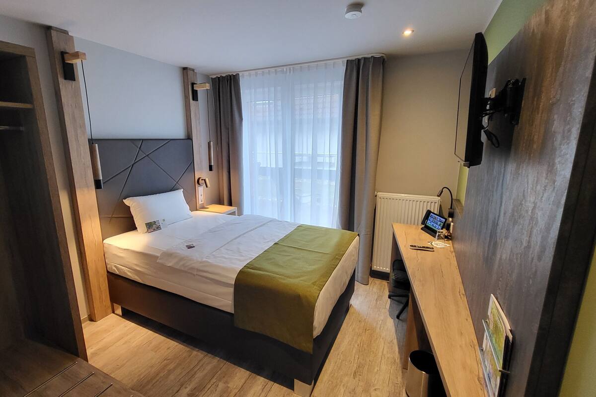 Ett hotellrum med en säng, ett nattduksbord med en tv, ett skrivbord med en dator och ett fönster med gardiner.