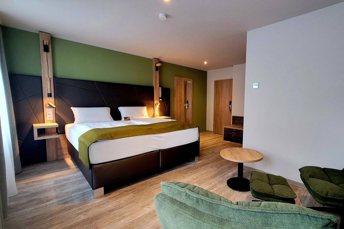 Ett modernt hotellrum med en stor säng, ett sängbord, en grön fåtölj och ett litet runt bord.