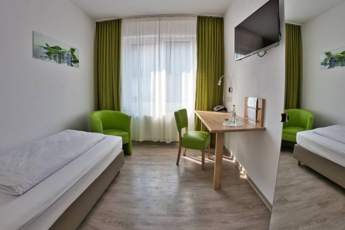 Ett hotellrum med två separata sängar, ett skrivbord med en stol, en platt-TV och ett fönster med tunna, genomskinliga gardiner.