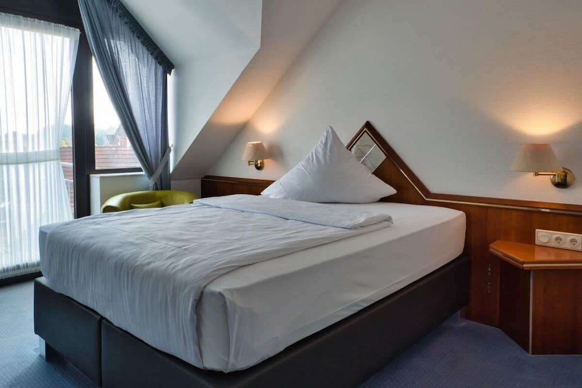 Ett modernt hotellrum med en stor säng, ett nattduksbord med en lampa och utsikt över omgivningarna genom ett fönster med gardiner.