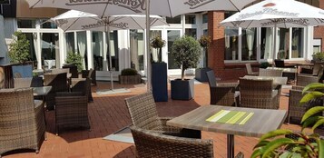 Un espacio al aire libre con mobiliario de mimbre y sombrillas, con suelo de ladrillo y plantas en macetas.