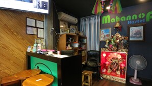 Une pièce comprenant un comptoir d’accueil, deux tabourets en bois, un fauteuil en cuir noir, un autel avec des objets religieux et un ventilateur.